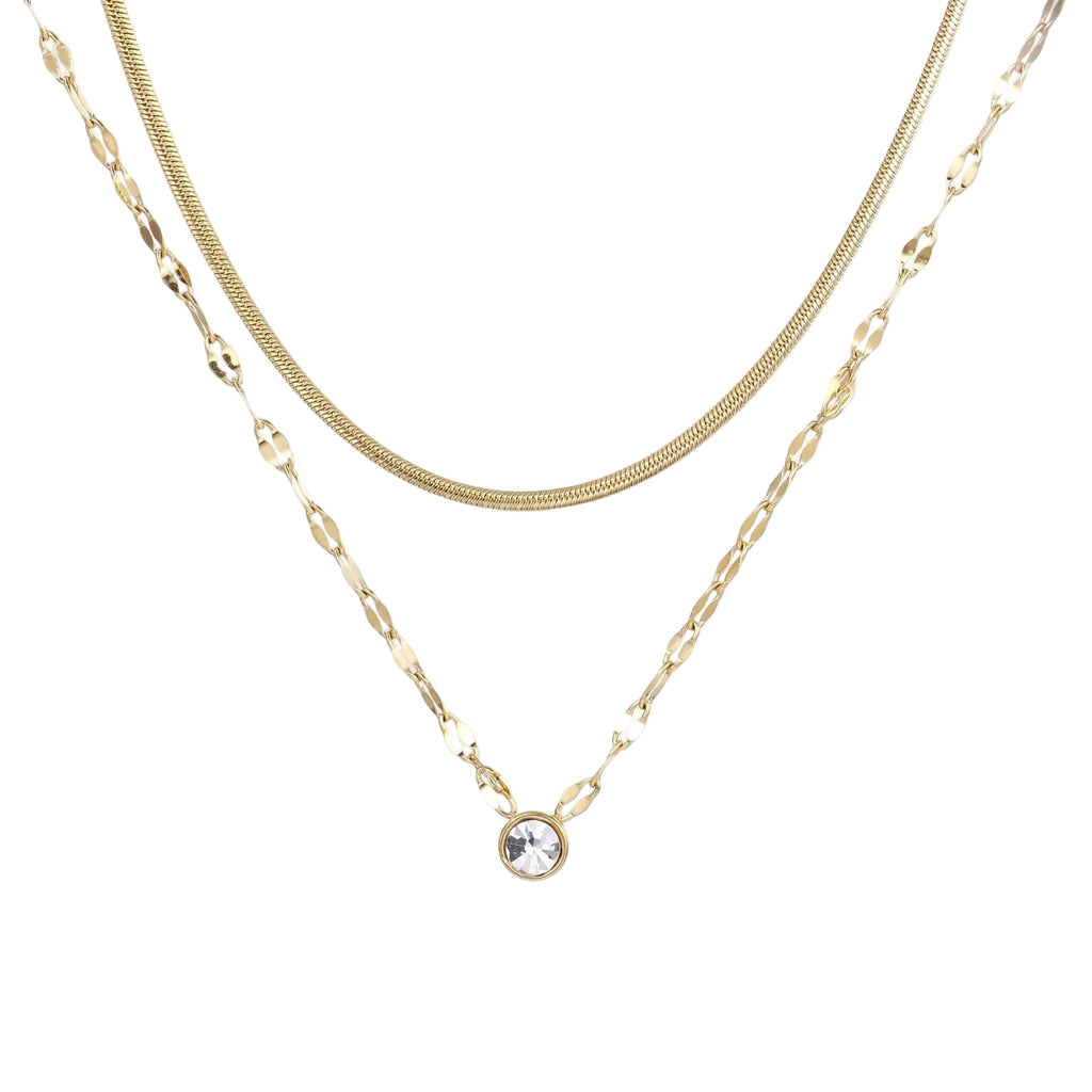Layered gold necklace with herringbone and link chains, small bezel-set crystal pendant on cream background