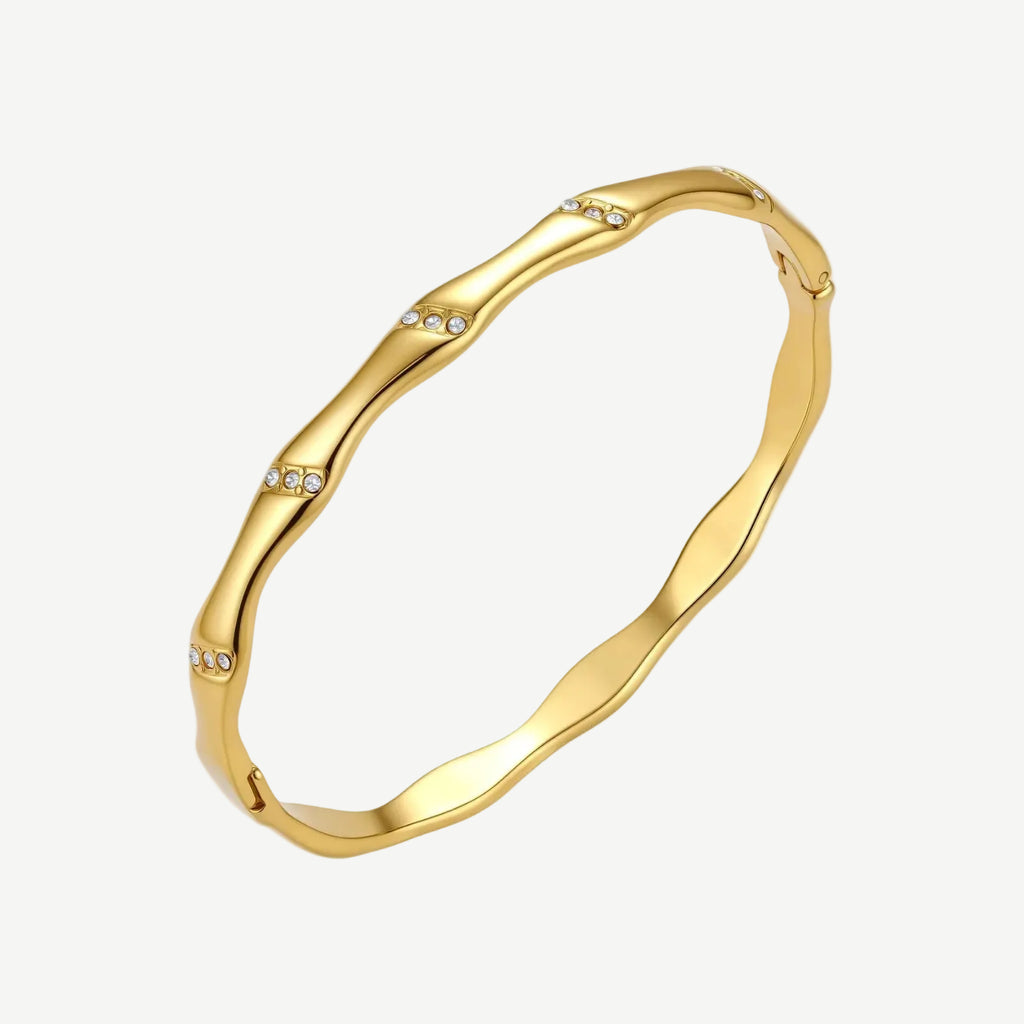 Gold-toned wavy bangle with bamboo-style segments and CZ crystal clusters on cream background. 18K plated stainless steel.