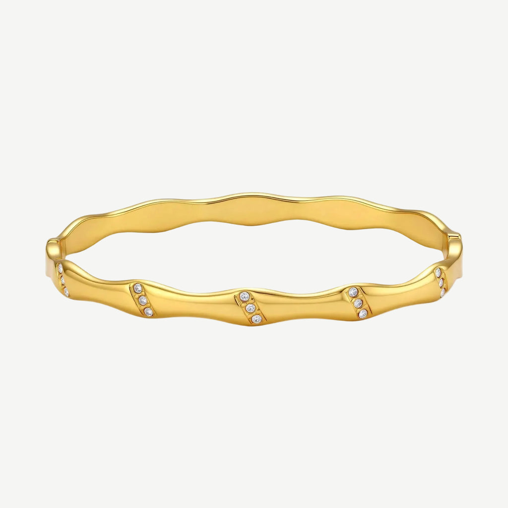 Gold-toned wavy bangle with bamboo-style segments and CZ crystal clusters on cream background. 18K plated stainless steel.